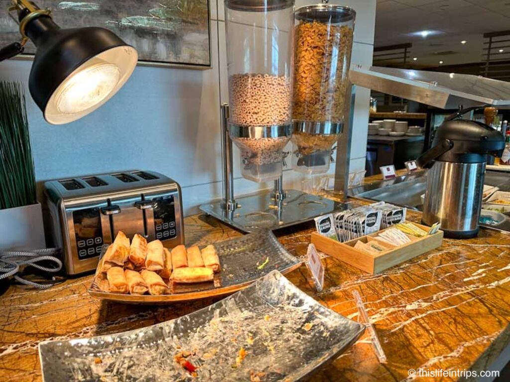Edmonton Plaza Premium lounge food under a heat lamp. Spring rolls next to a dry cereal dispenser.